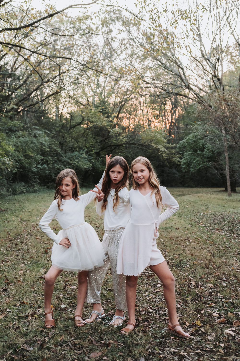 Three young friends in white dresses pose playfully together in a natural outdoor setting.