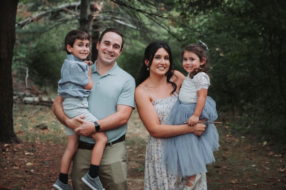 A family of four poses together for a portrait in a wooded setting at dusk.