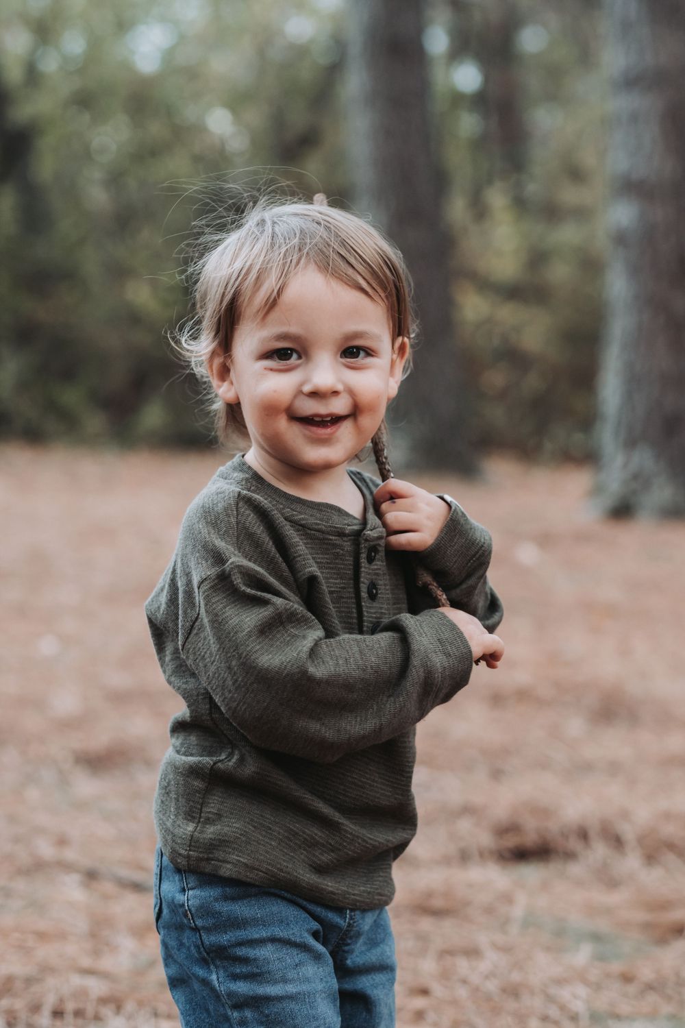 Toddler in green sweater smiling while playing in wooded area.