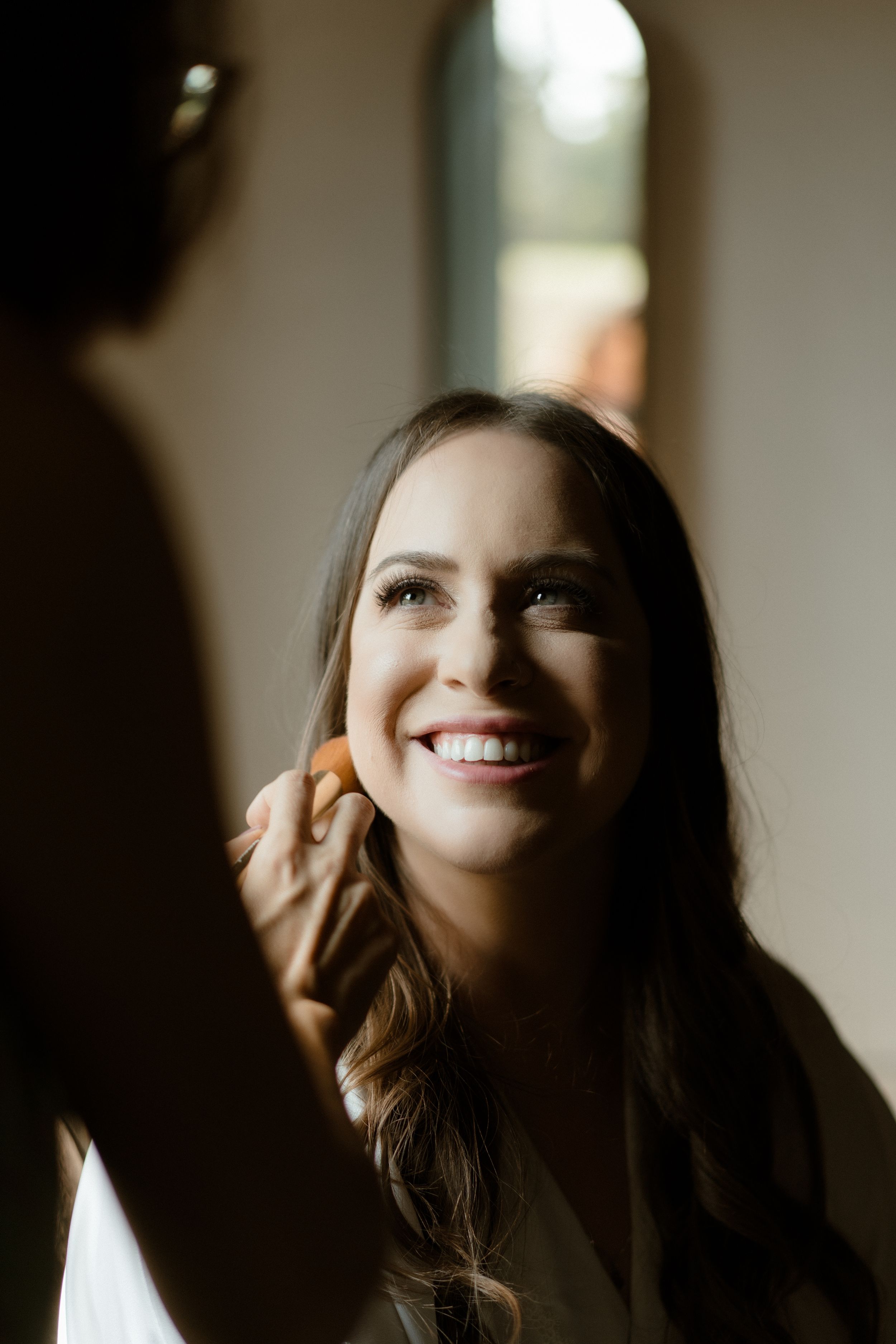 Soft natural window light illuminates wedding preparation scene.
