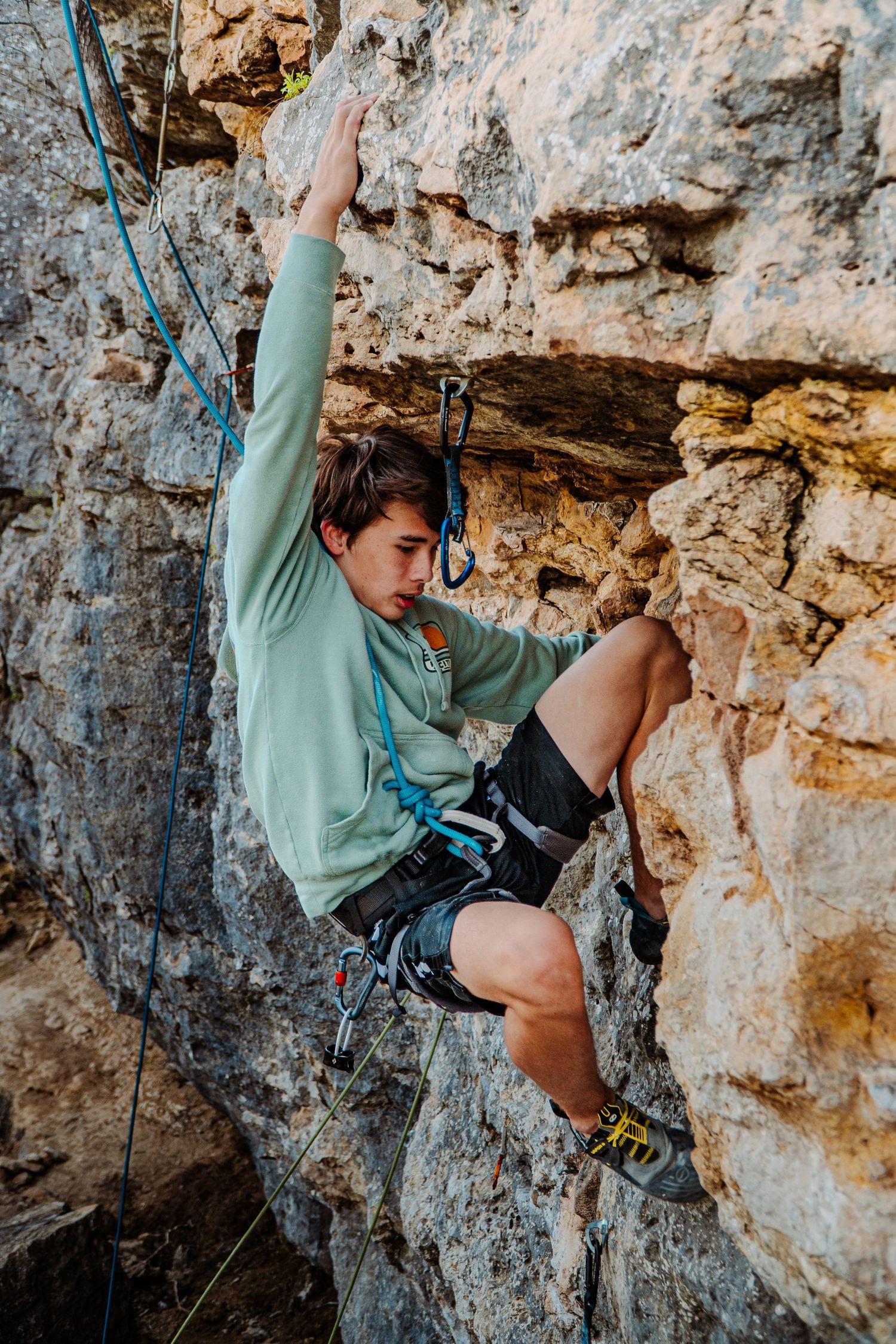 A climber in mint green hoodie scales a challenging limestone rock face with proper safety gear.