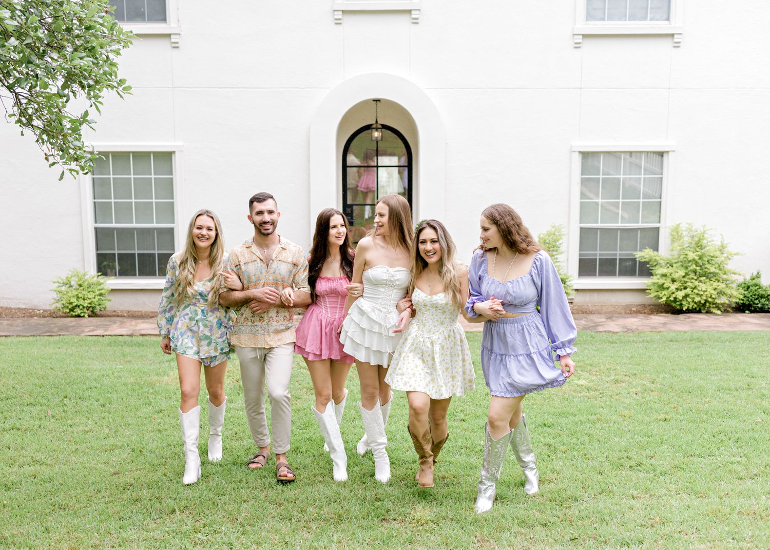 Group in pastel dresses and white boots pose in front of a white historic building.