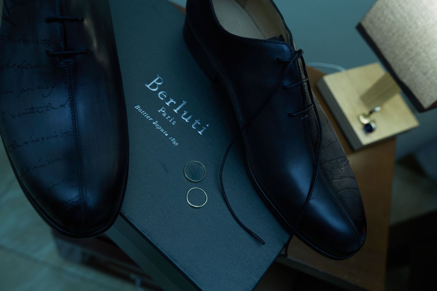 Black formal shoes displayed next to a small table lamp on a dark surface.