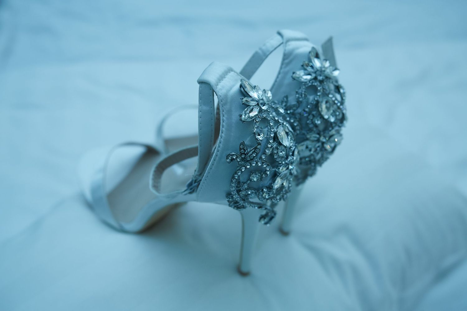 Elegant white bridal heels with crystal embellishments shown against a light blue background.