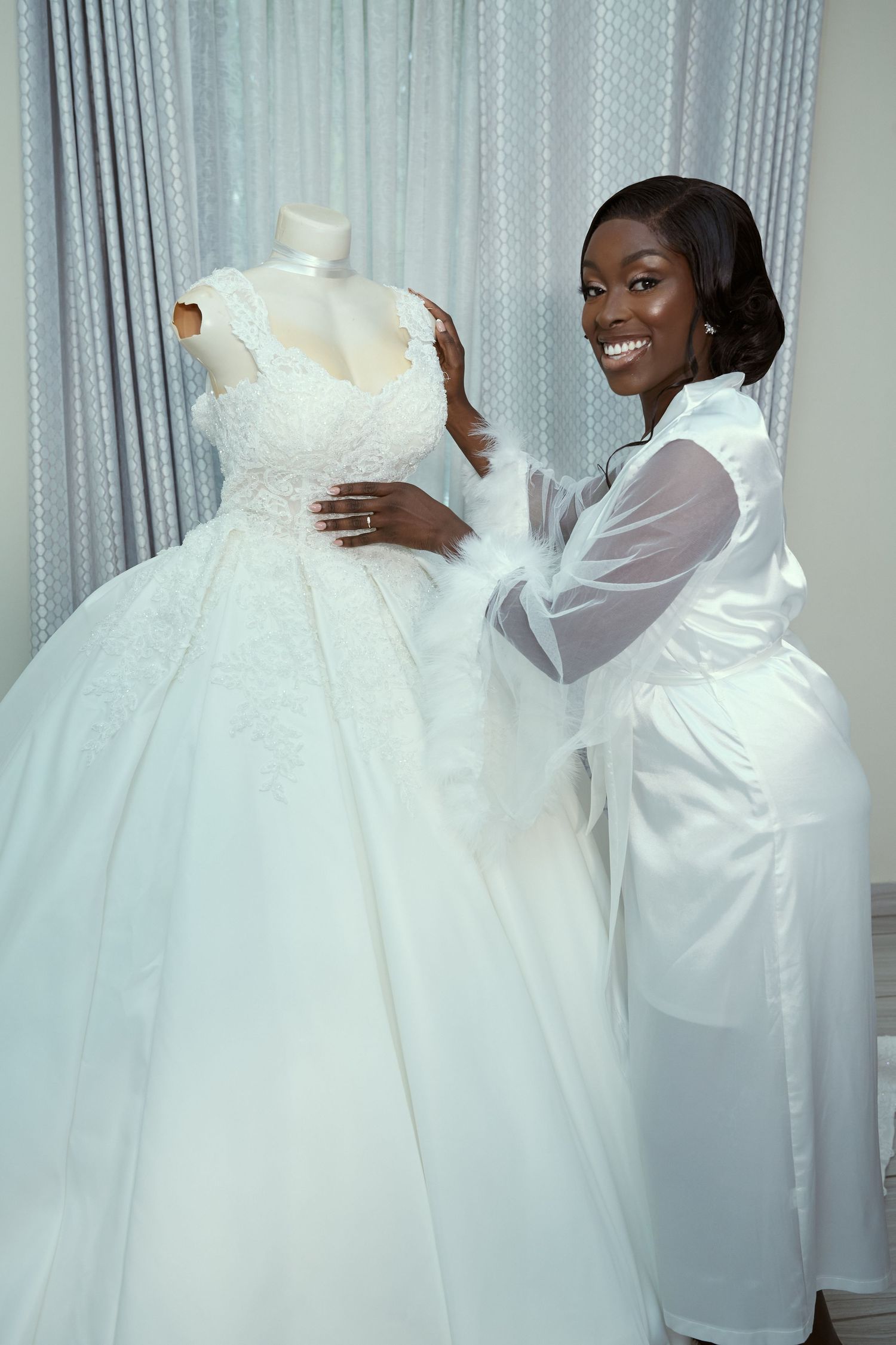A flowing white wedding gown being displayed against light curtains in a bridal fitting room.