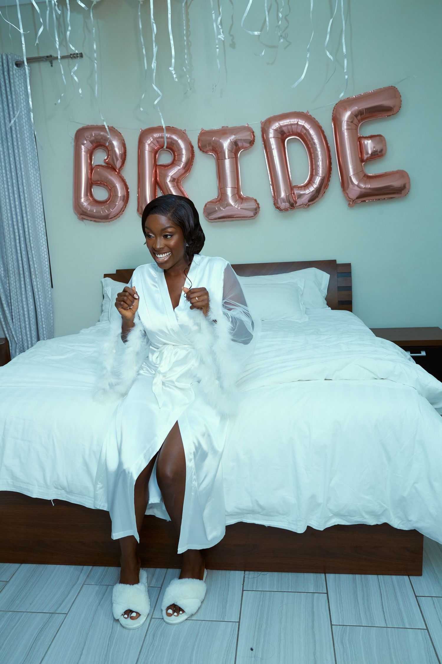 Bridal preparation scene with white robe and decorative balloon letters in the background.