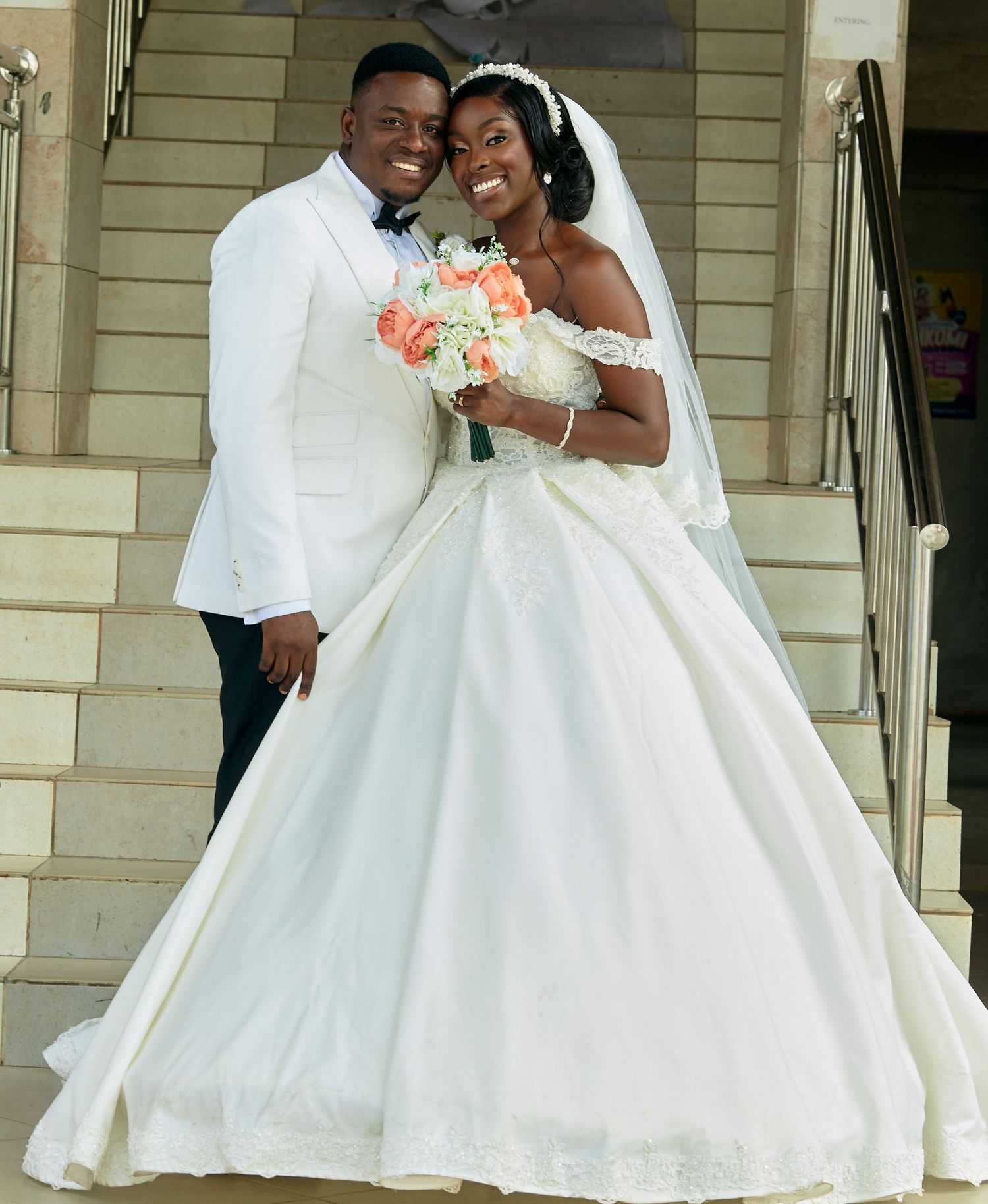 Wedding couple poses on stone steps in elegant white attire with floral bouquet.