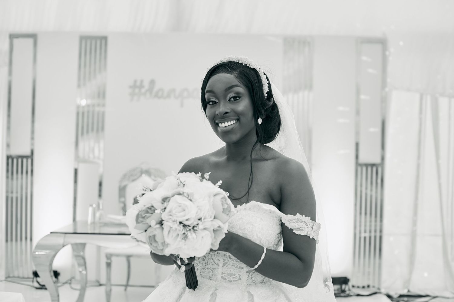 A joyful bride in an off-shoulder wedding dress holds a white bouquet while smiling radiantly in black and white.