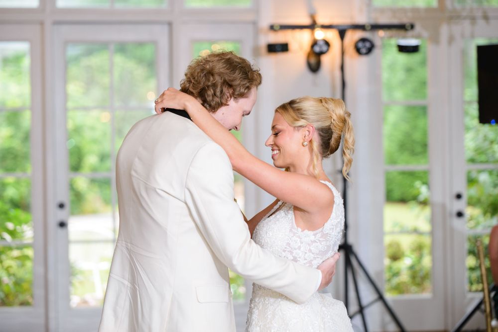 A couple shares their first dance at a wedding reception with bright windows and lighting equipment in the background.