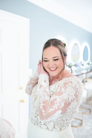 A person in an elegant white lace wedding gown smiles joyfully while touching their hair in a bright room. primrose cottage photos