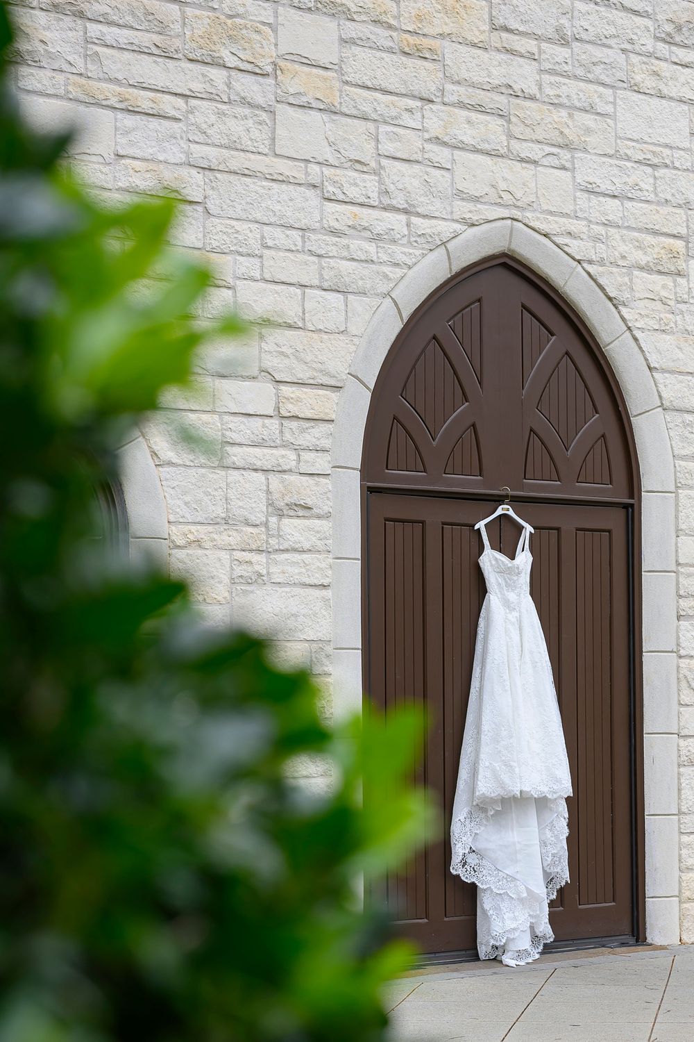 White lace wedding dress hanging on arched brown church door with stone wall backdrop. Ashton Gardens Wedding Photos