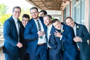 A group of groomsmen in matching navy blue suits smile and huddle together under a covered porch.