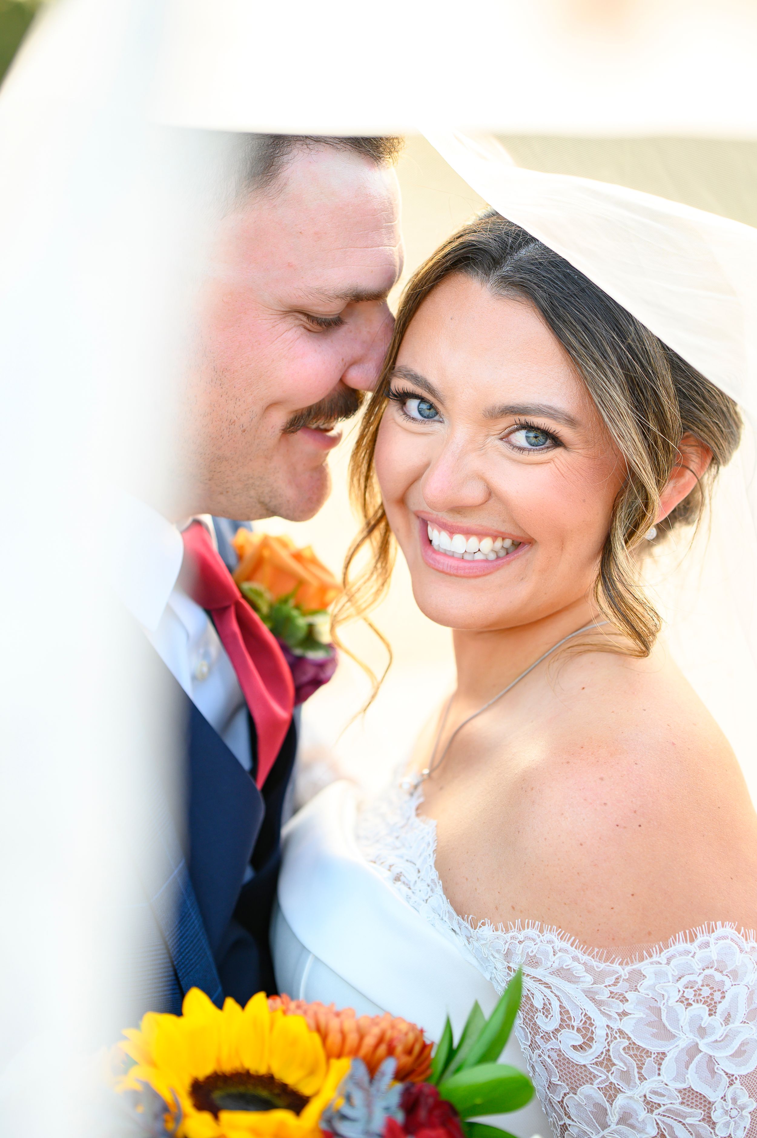 Smiling bride in lace dress with veil holds sunflower bouquet beside groom in navy suit with coral tie.