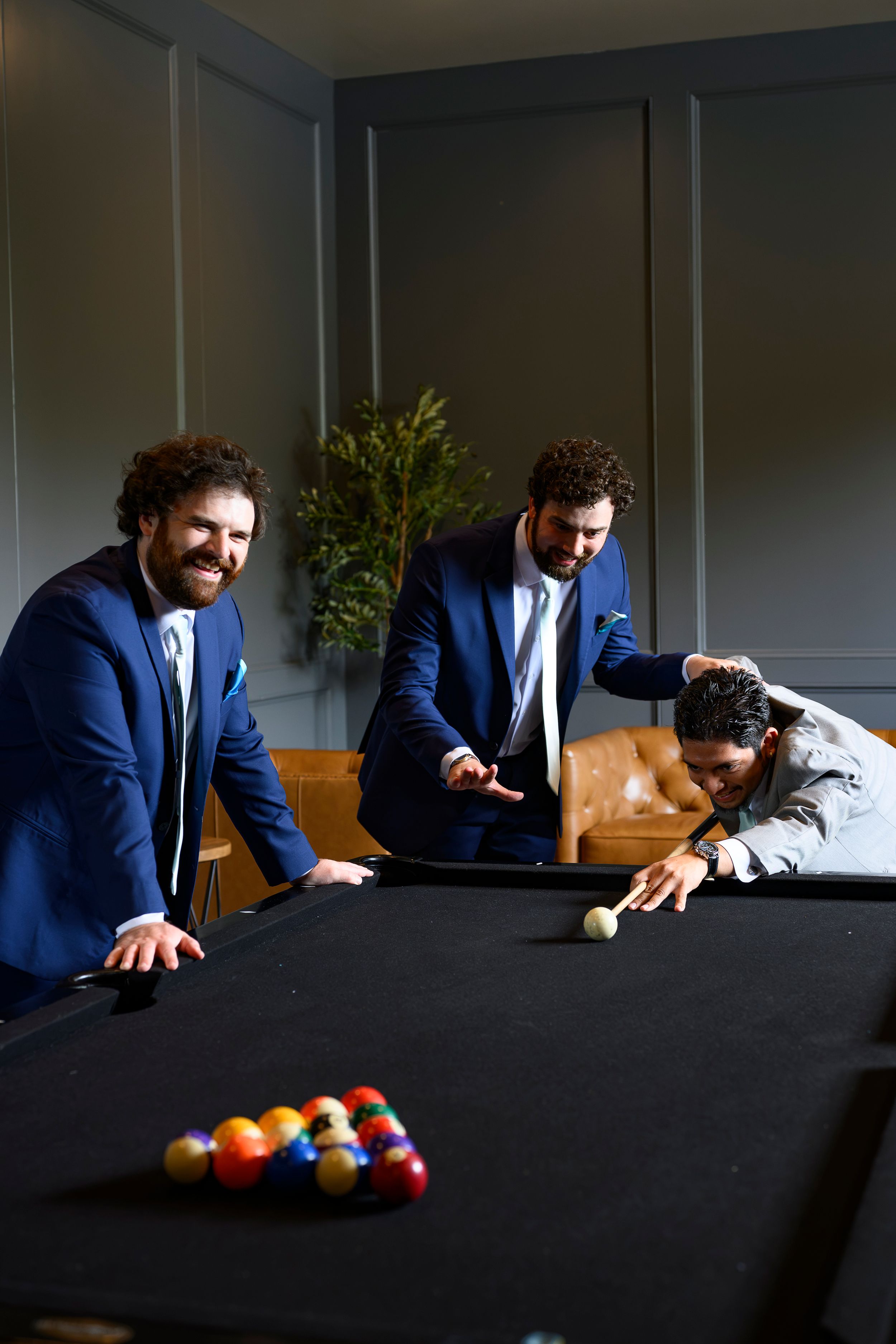 Three men in blue suits playing pool billiards, one leaning over black table to take a shot.