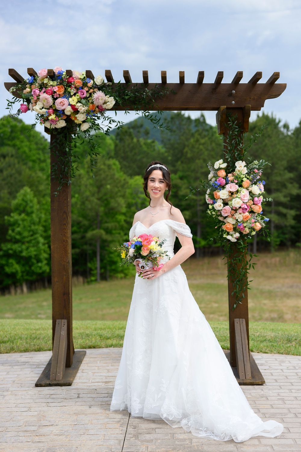 Bride in white off-shoulder gown holding colorful bouquet under floral wooden arch at outdoor wedding venue.