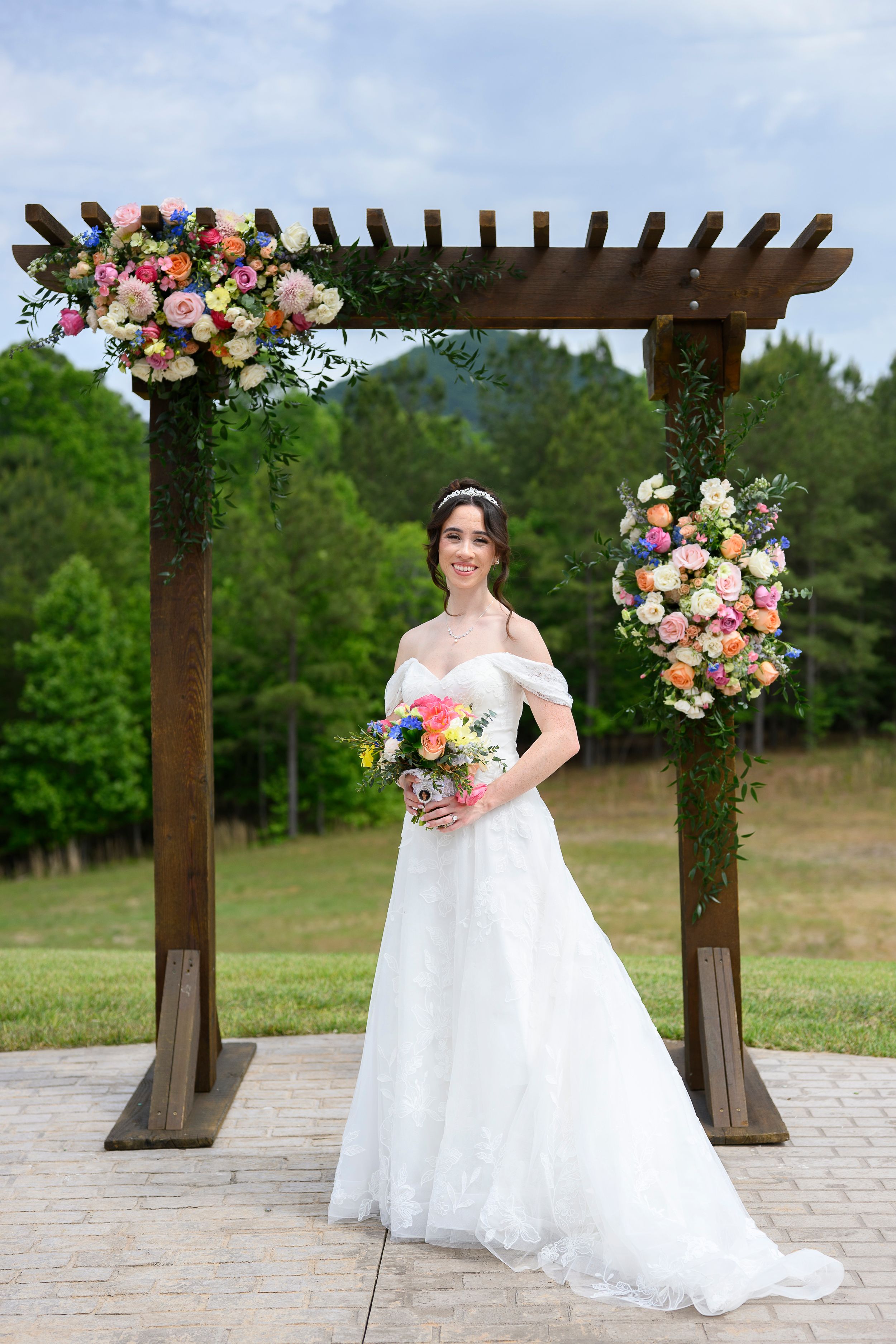 Bride in white off-shoulder gown holding colorful bouquet under floral wooden arch at outdoor wedding venue.