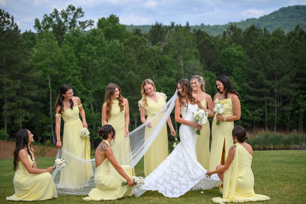 Bride in white gown with bridesmaids in yellow dresses posing outdoors with green trees and mountains backdrop.