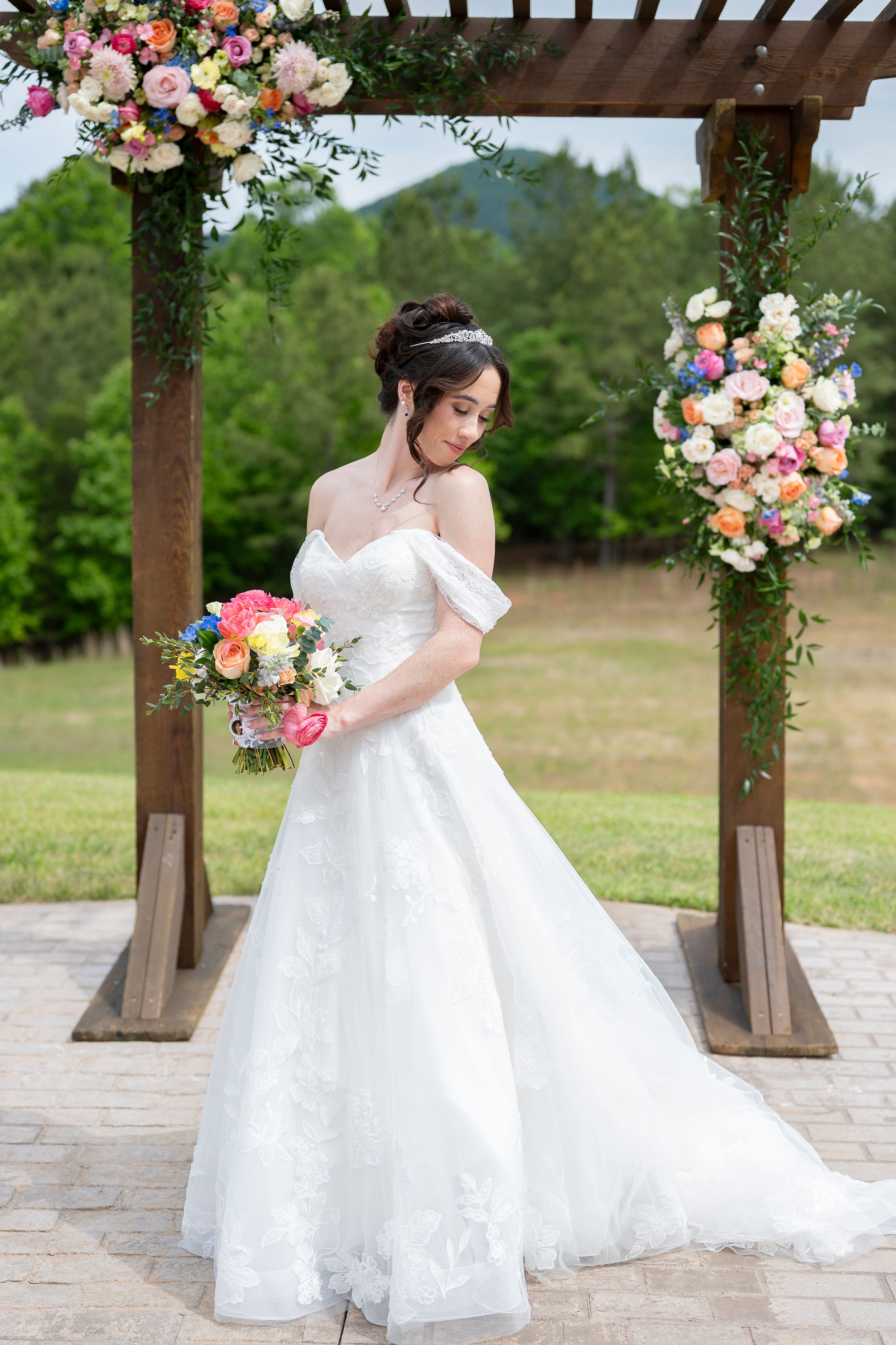 Bride in off-shoulder white ball gown holds colorful bouquet under floral pergola at outdoor wedding ceremony.