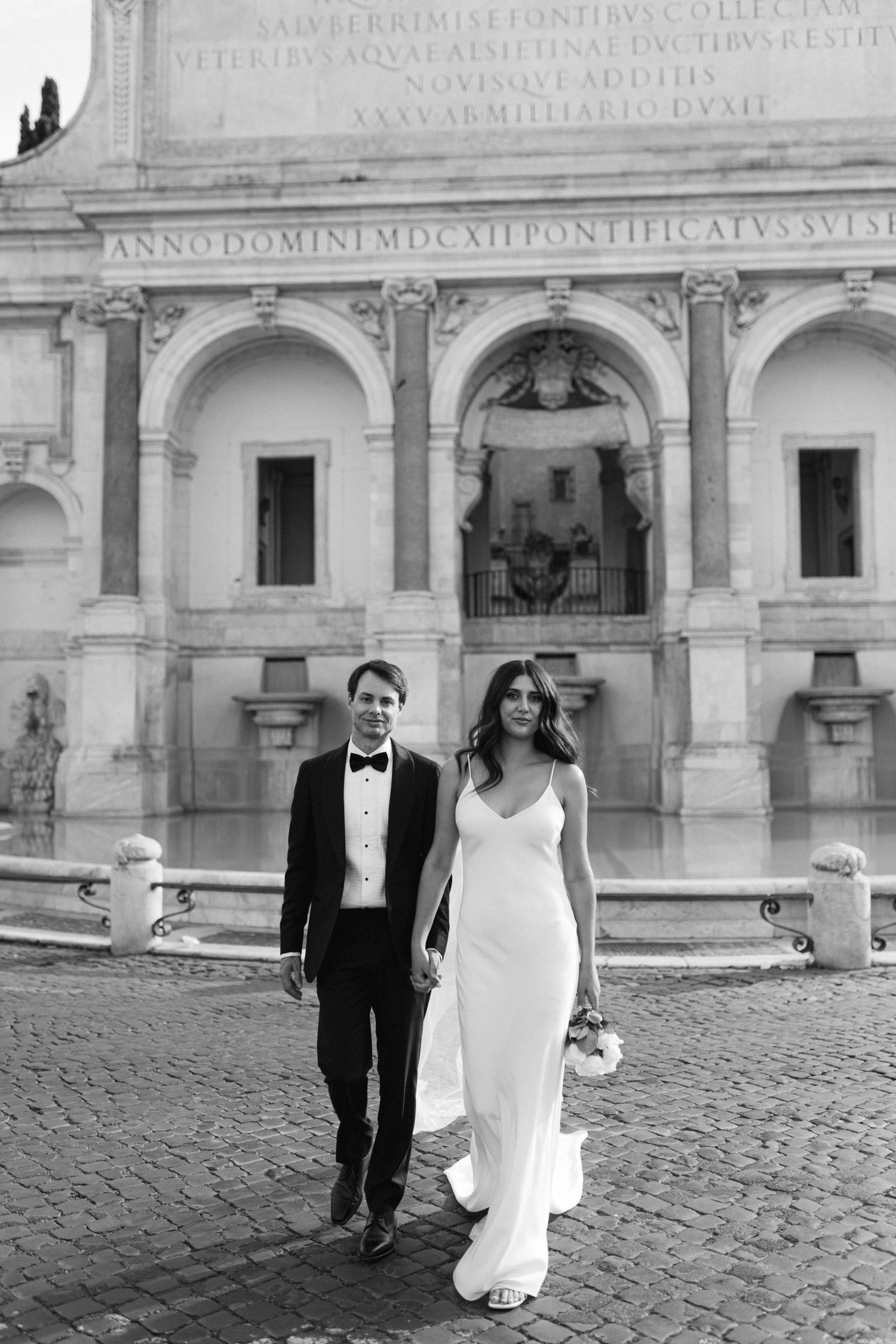 Elegant couple in formal wedding attire walk along cobblestones in front of historic Italian building with arched architecture in Rome.
