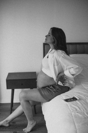 A black and white maternity photo showing a pregnant woman sitting on a bed.