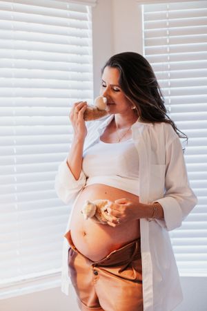 A pregnant woman in white clothing stands near window blinds eating a snack.
