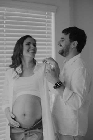 An intimate maternity photoshoot featuring a couple in white shirts by a window with blinds.