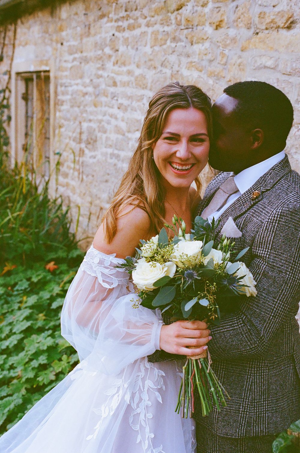 A romantic wedding scene with a couple embracing outdoors against a stone wall backdrop with white floral bouquet.