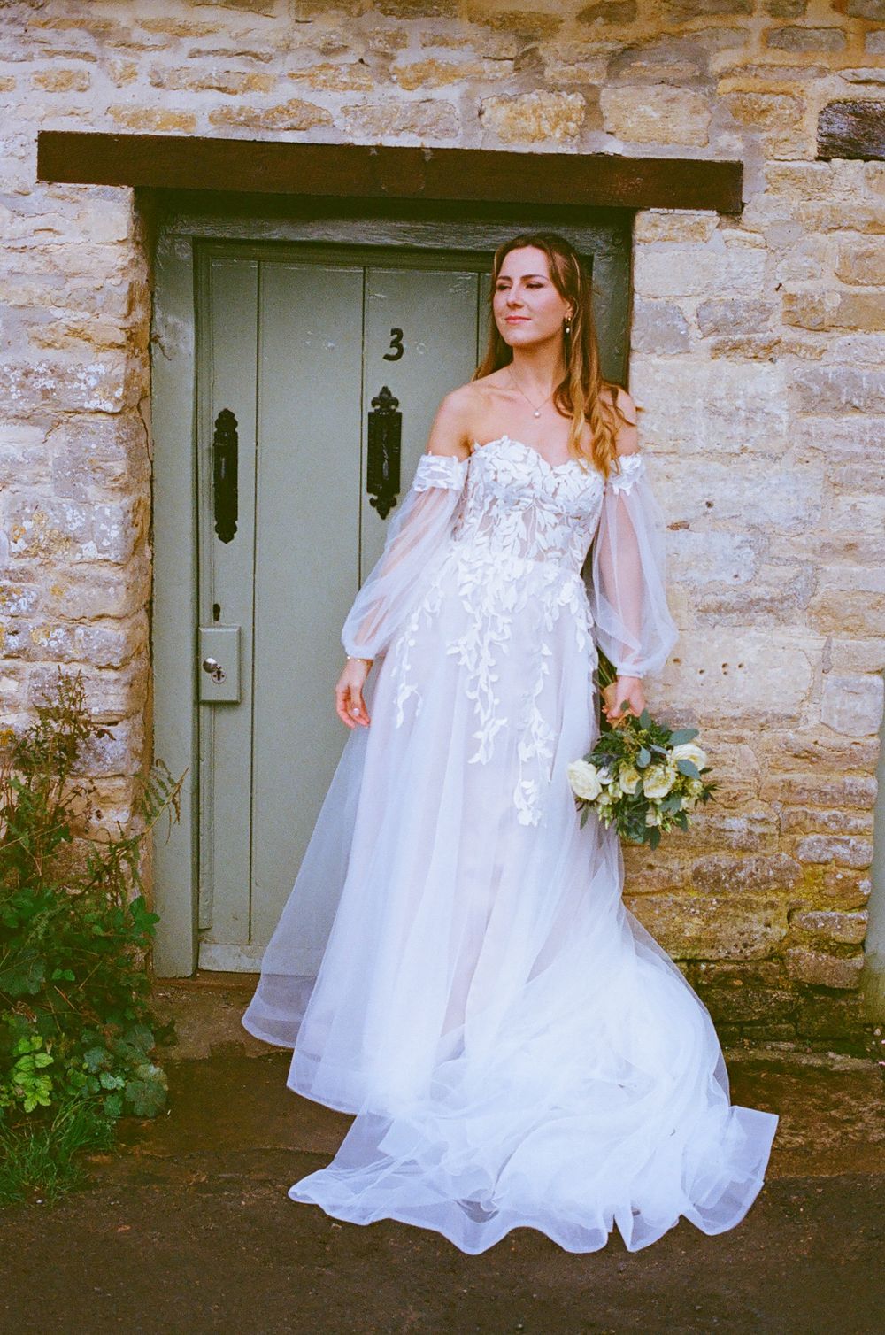 An elegant bride in a white lace gown poses in front of a rustic stone wall with a sage green door.