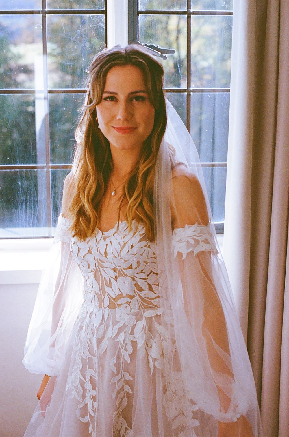 A sunlit indoor bridal portrait showcasing an intricate lace wedding gown with flowing sleeves.