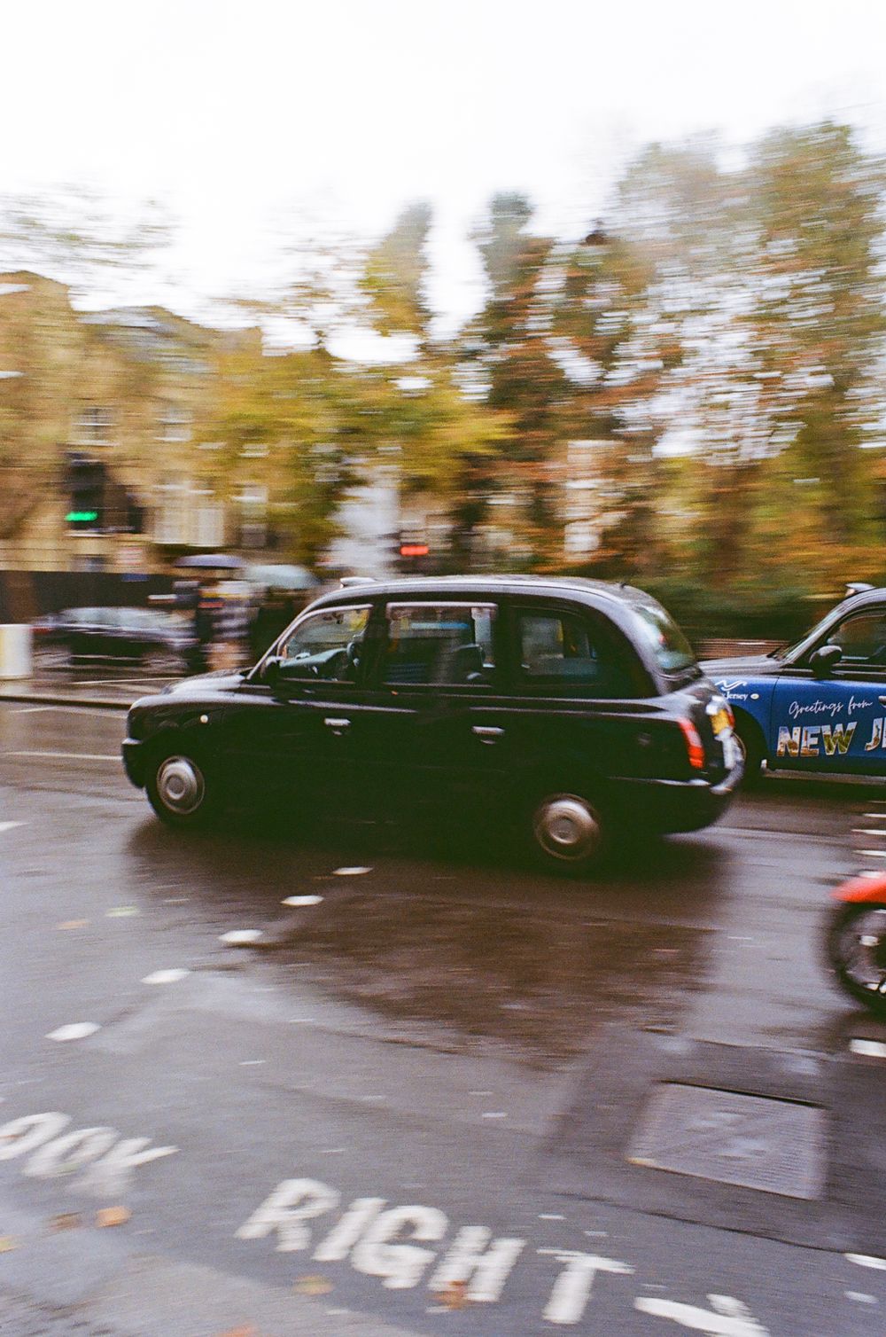 Vintage black cars driving in a row on a rainy street with autumn trees in the background.