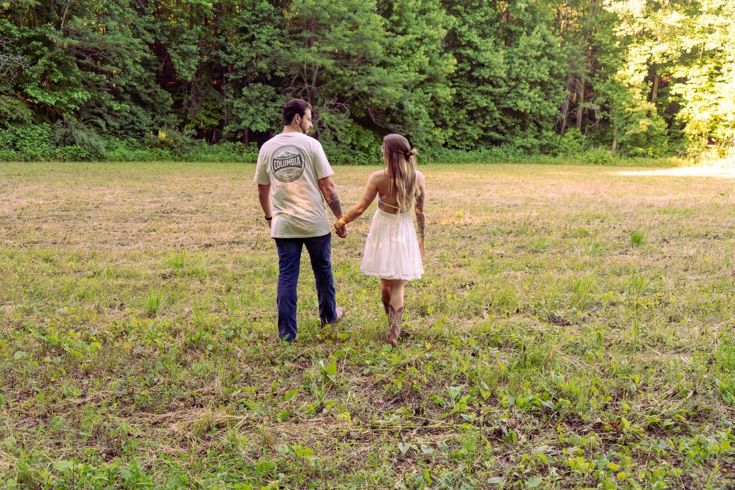 Couple holding hands walking through a grassy field surrounded by trees.