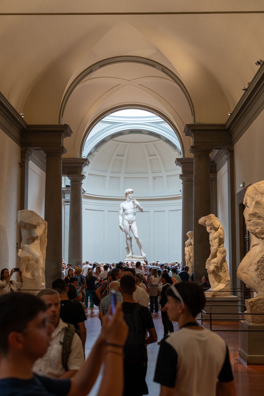 Michelangelo's David statue displayed in crowded museum gallery under domed ceiling.