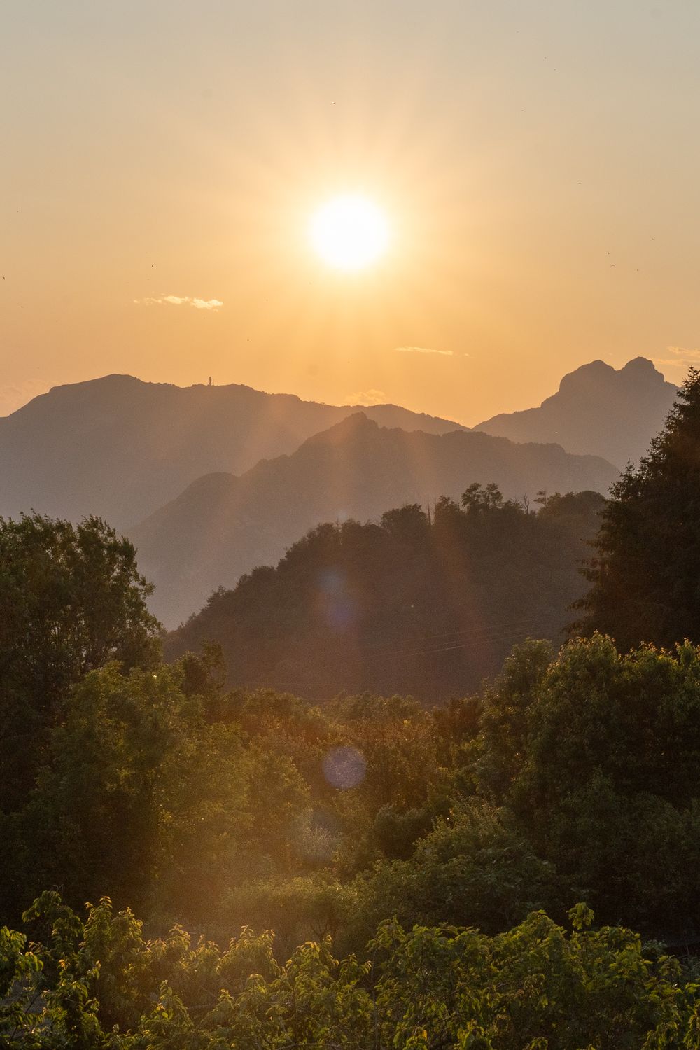 Golden sun sets behind mountain silhouettes creating a warm evening glow over forested hills.