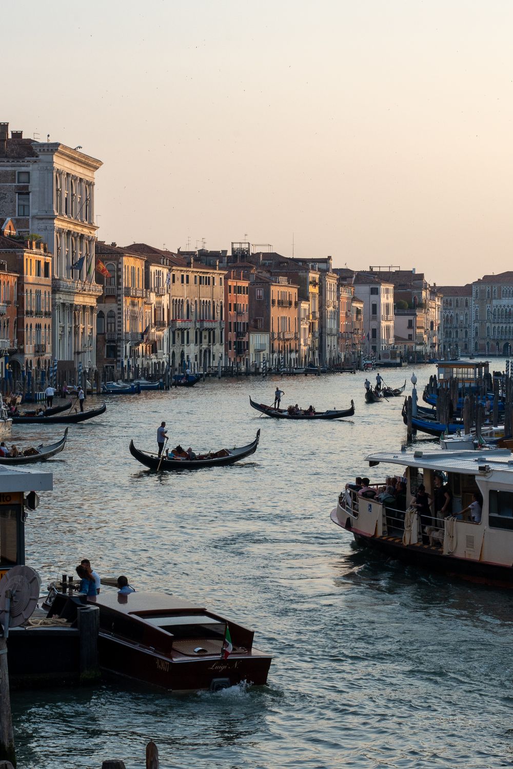 Gondolas navigate the Grand Canal at sunset among historic Venetian buildings and water traffic.