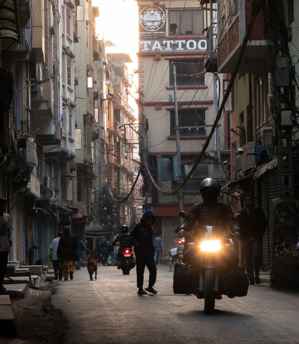 Narrow alley in Kathmandu with a tattoo shop sign and motorcycle headlight glowing at dusk.
