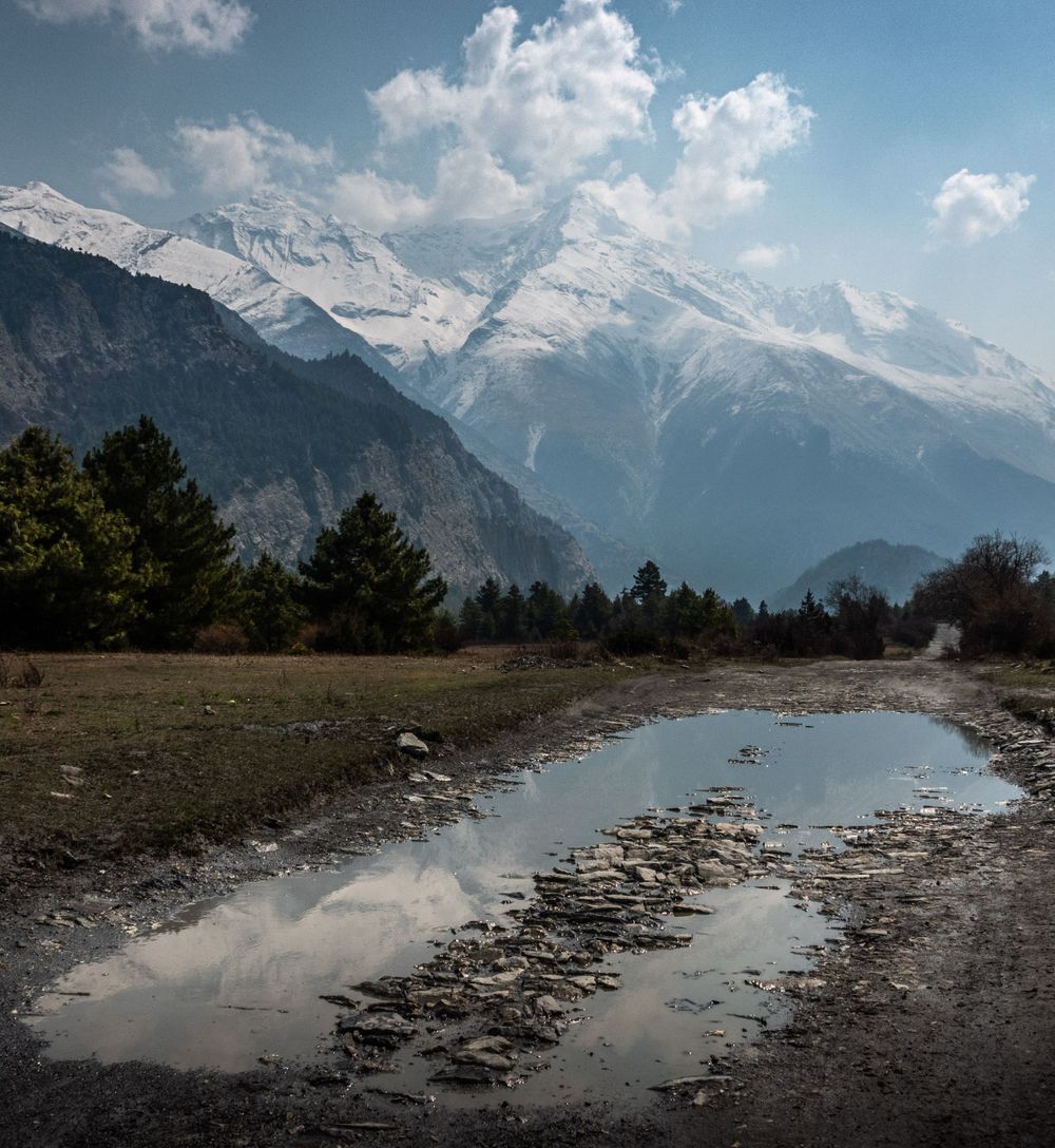 Snow-capped Himalayan mountains reflected in a muddy puddle with blue sky and clouds.