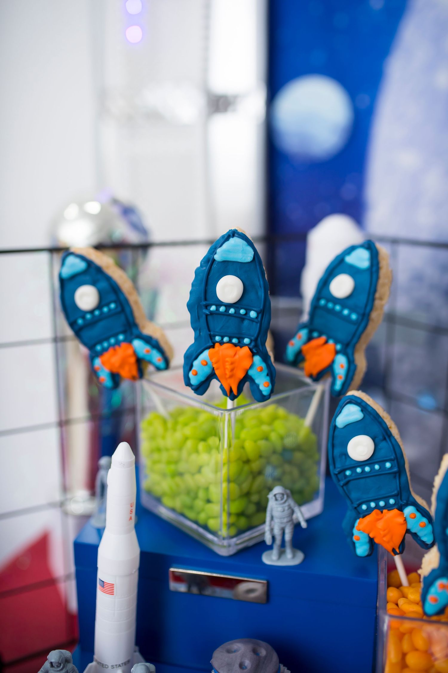 Blue rocket ship cookies displayed in clear container with space-themed decorations.