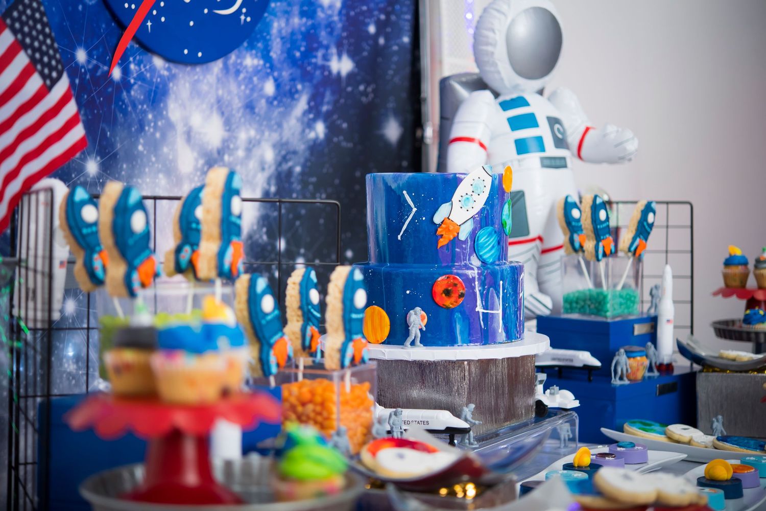 Space-themed party dessert table with rocket cake, American flag, and blue decorations.