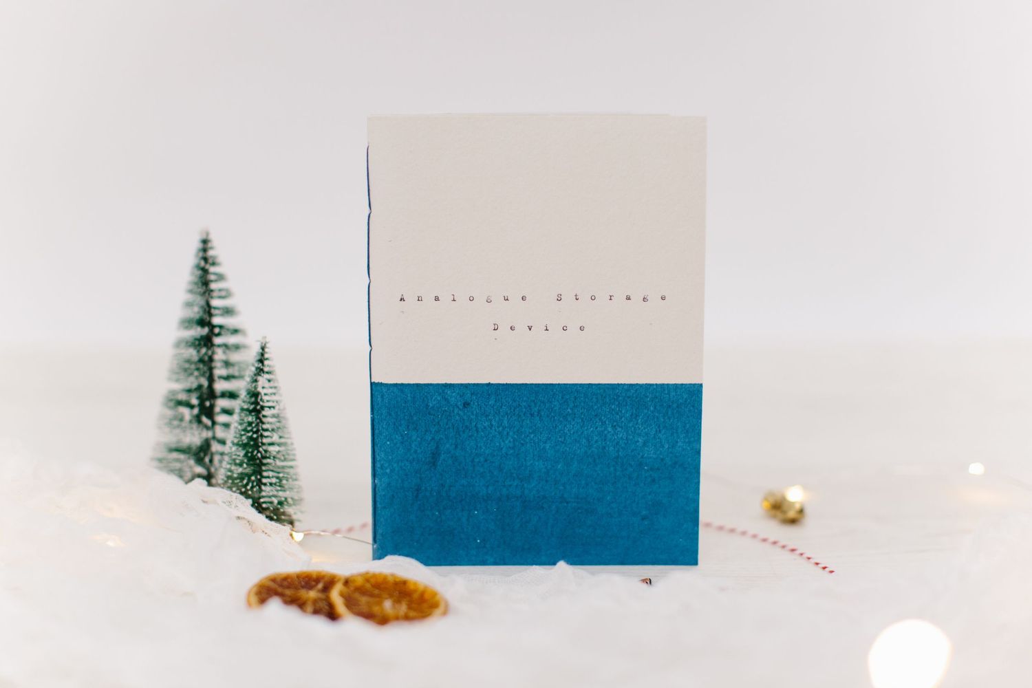 A blue and white greeting card displayed in snow with evergreen trees in a soft winter scene background.