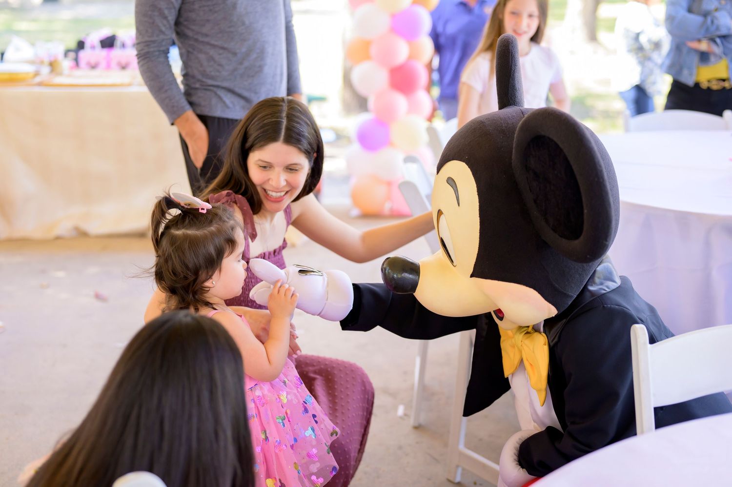 Mickey Mouse character greets guests at a festive party with pink balloon decorations and party decor.