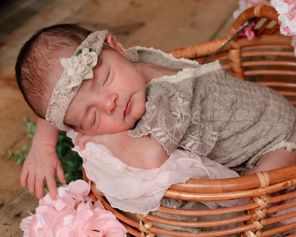 Sleeping infant in lace outfit resting in wicker basket with pink floral decor.