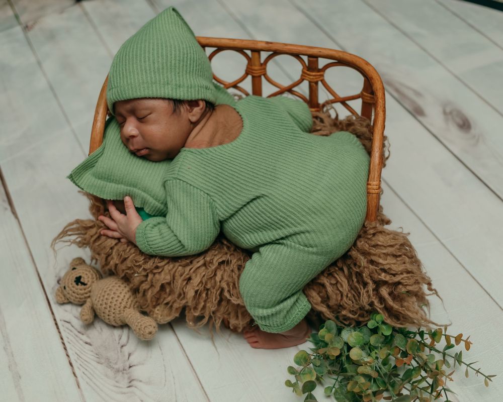 Sleeping infant in green knit outfit and hat lying in rattan chair with decorative accents.