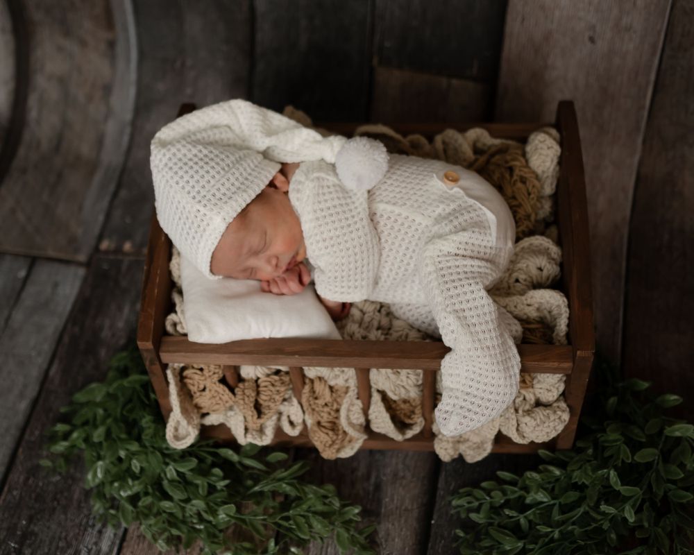Newborn baby sleeping peacefully in white knit outfit on rustic wooden prop bed.