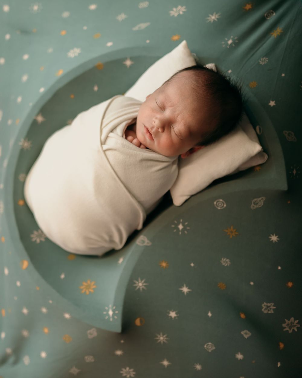 Swaddled newborn sleeping on starry teal background with celestial pattern.
