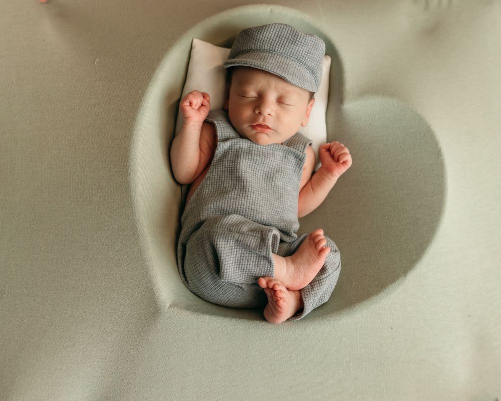 Sleeping newborn in grey outfit stretching arms while resting in cream-colored chair.
