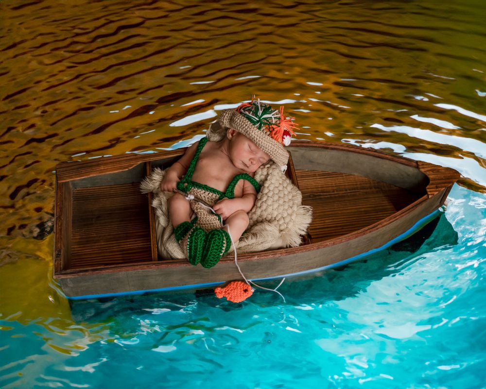 Newborn baby in green outfit sleeping in wooden boat prop on blue and brown water for creative photoshoot.