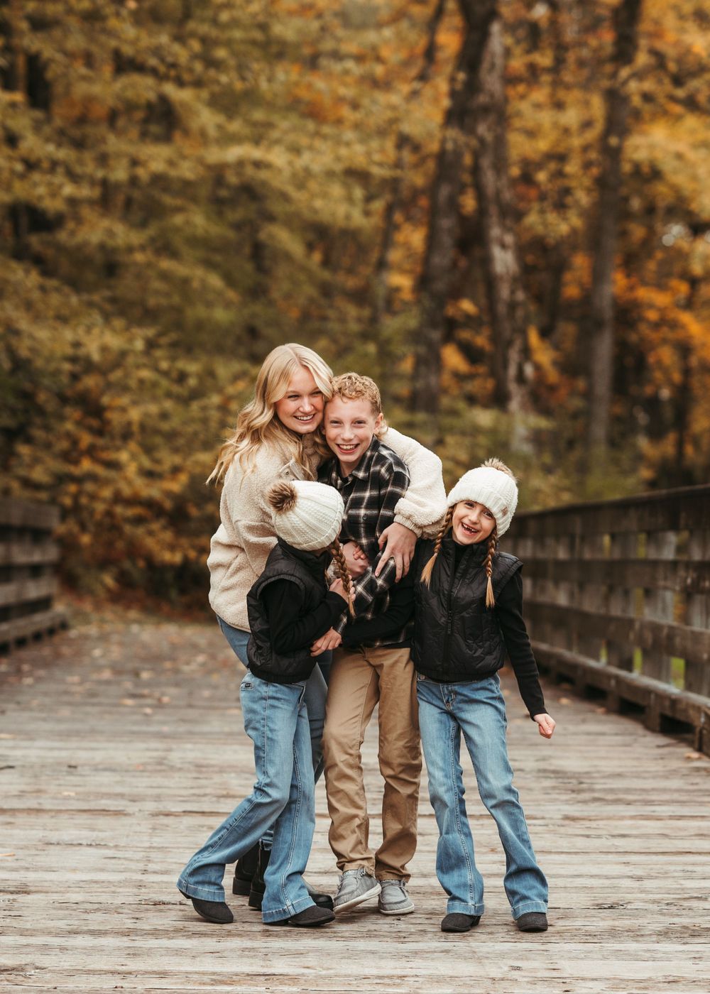 Three people embracing and laughing on wooden bridge surrounded by fall foliage.