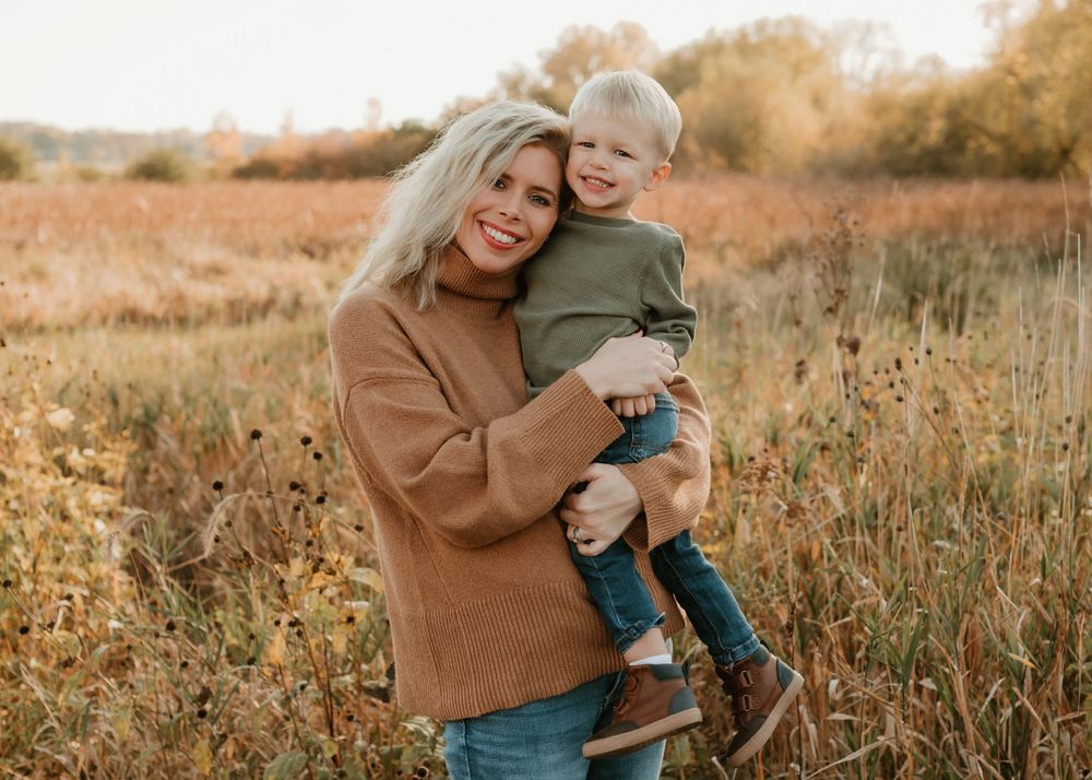Person in tan sweater embracing small child in casual wear against autumn backdrop.