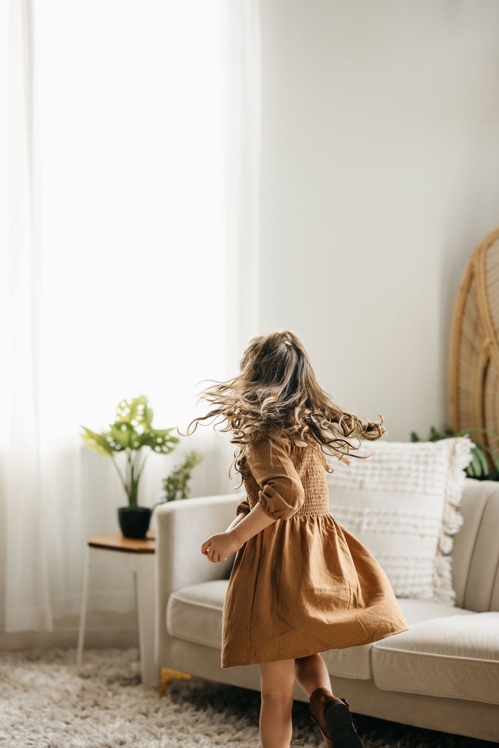 Child in brown dress twirls playfully in bright minimalist living room with white sofa and potted plants.