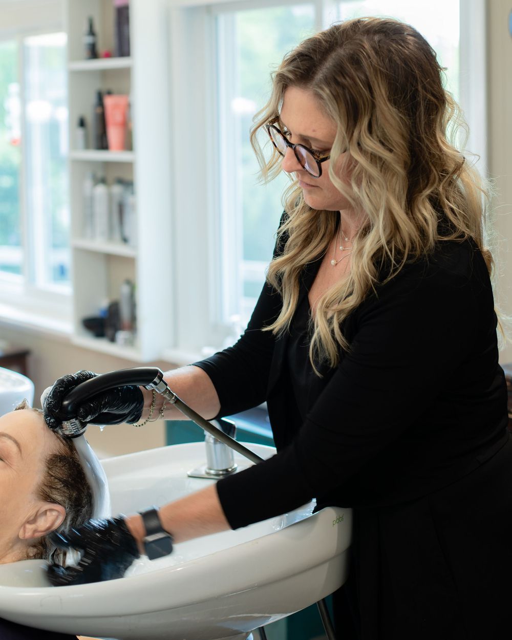 Stylist washing client's hair at salon sink basin wearing black gloves.